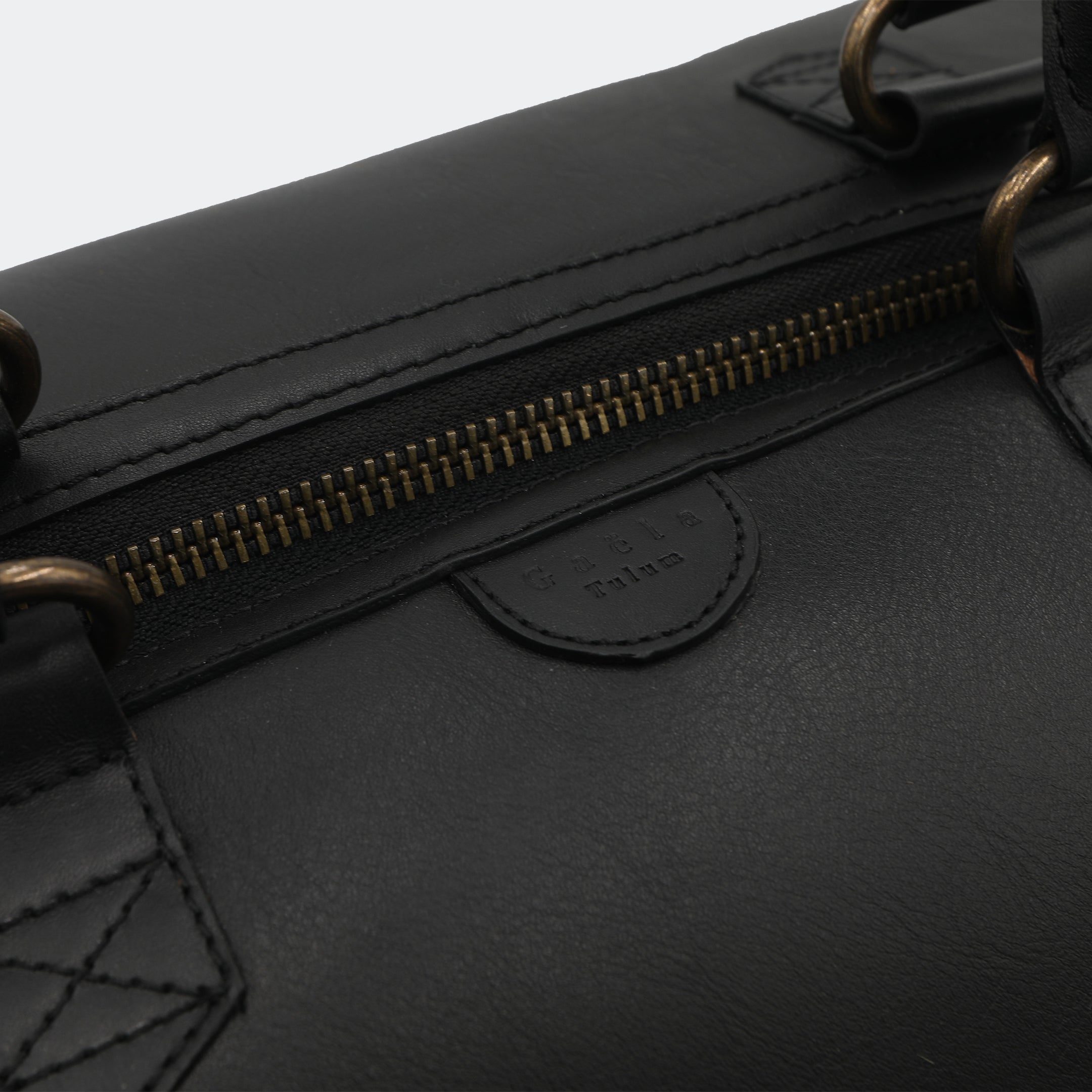 Close-up of The Valise in black leather, showing the curved leather handles, premium hardware, and refined grain texture of the leather.