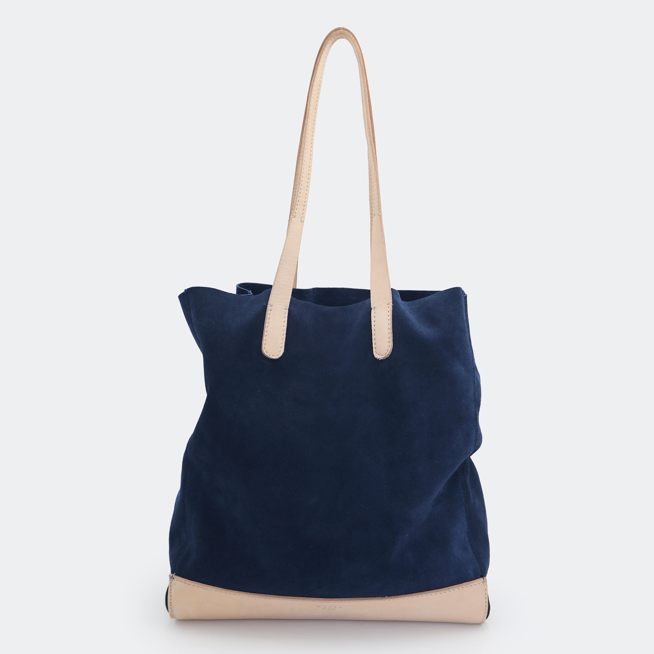 The Tote Bag in navy blue suede with full-grain leather accents on the base and handles, featuring a minimalist silhouette and structured shape.