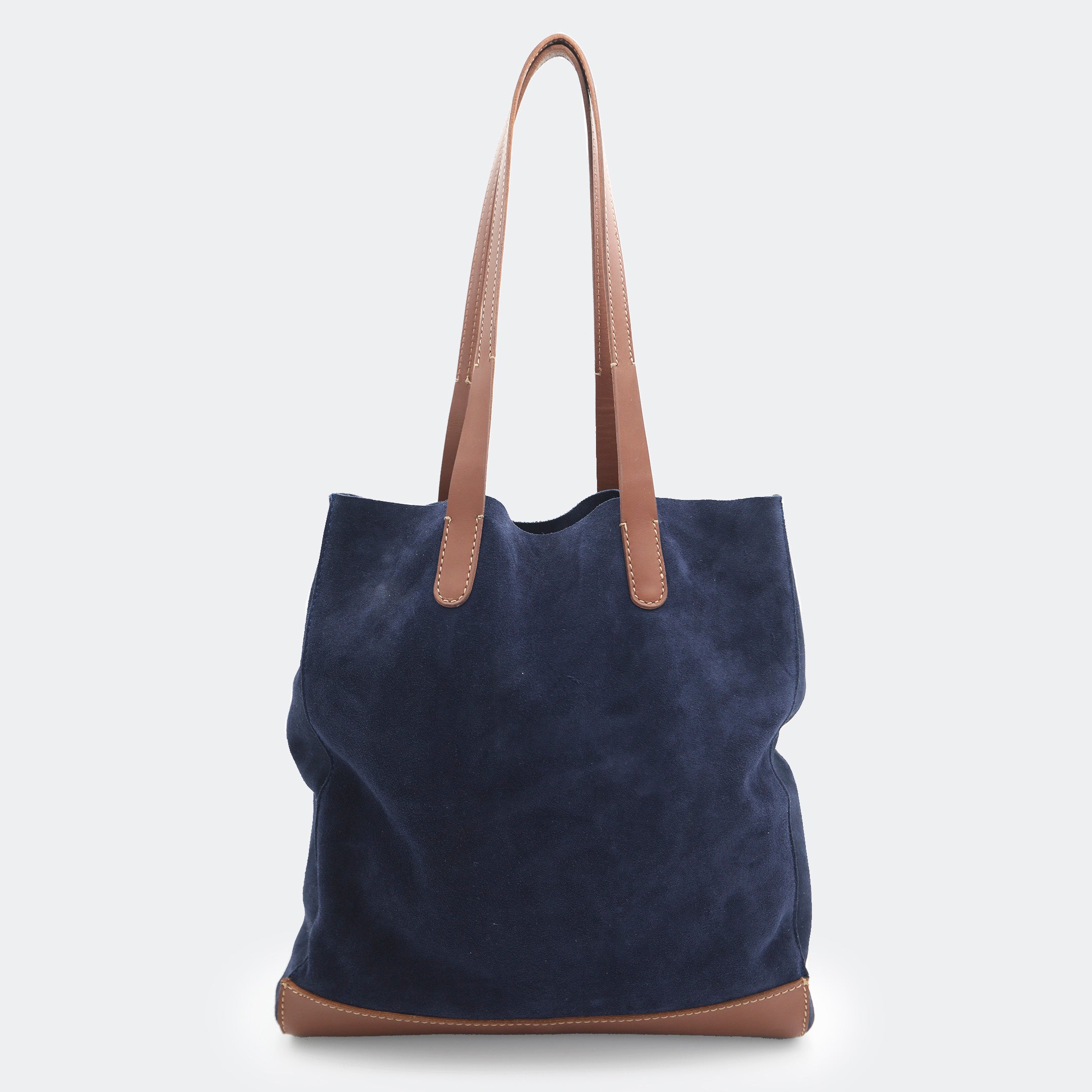 The Tote Bag in navy blue suede with brown full-grain leather accents on the base and handles, featuring a minimalist silhouette and structured shape.