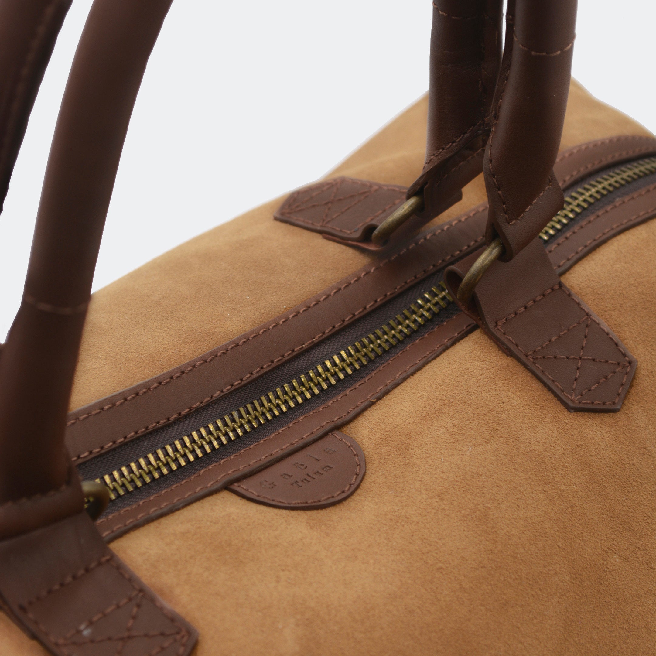Close-up of The Valise in brown leather, showing the curved leather handles, premium hardware, and refined grain texture of the leather.