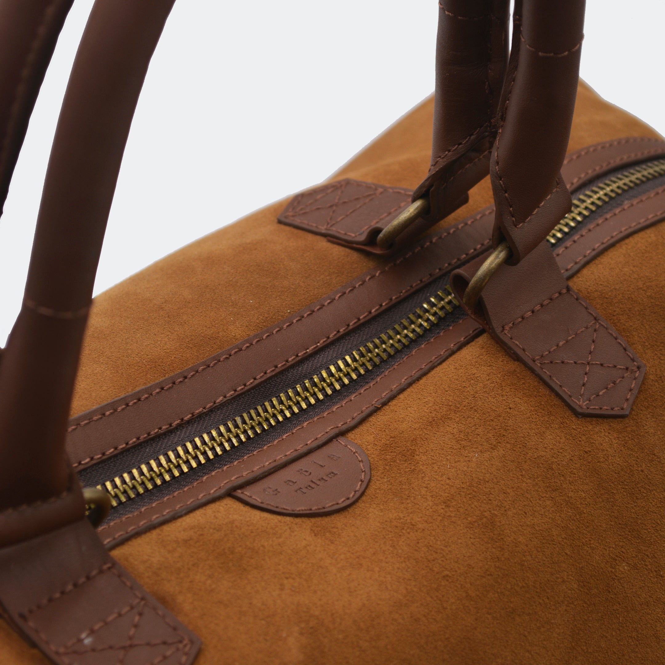 Close-up of The Valise in brown leather, showing the curved leather handles, premium hardware, and refined grain texture of the leather.