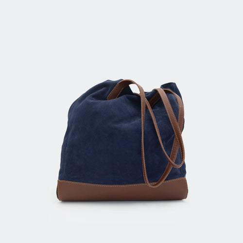 The Tote Bag in navy blue suede with brown full-grain leather accents on the base and handles, featuring a minimalist silhouette and structured shape.