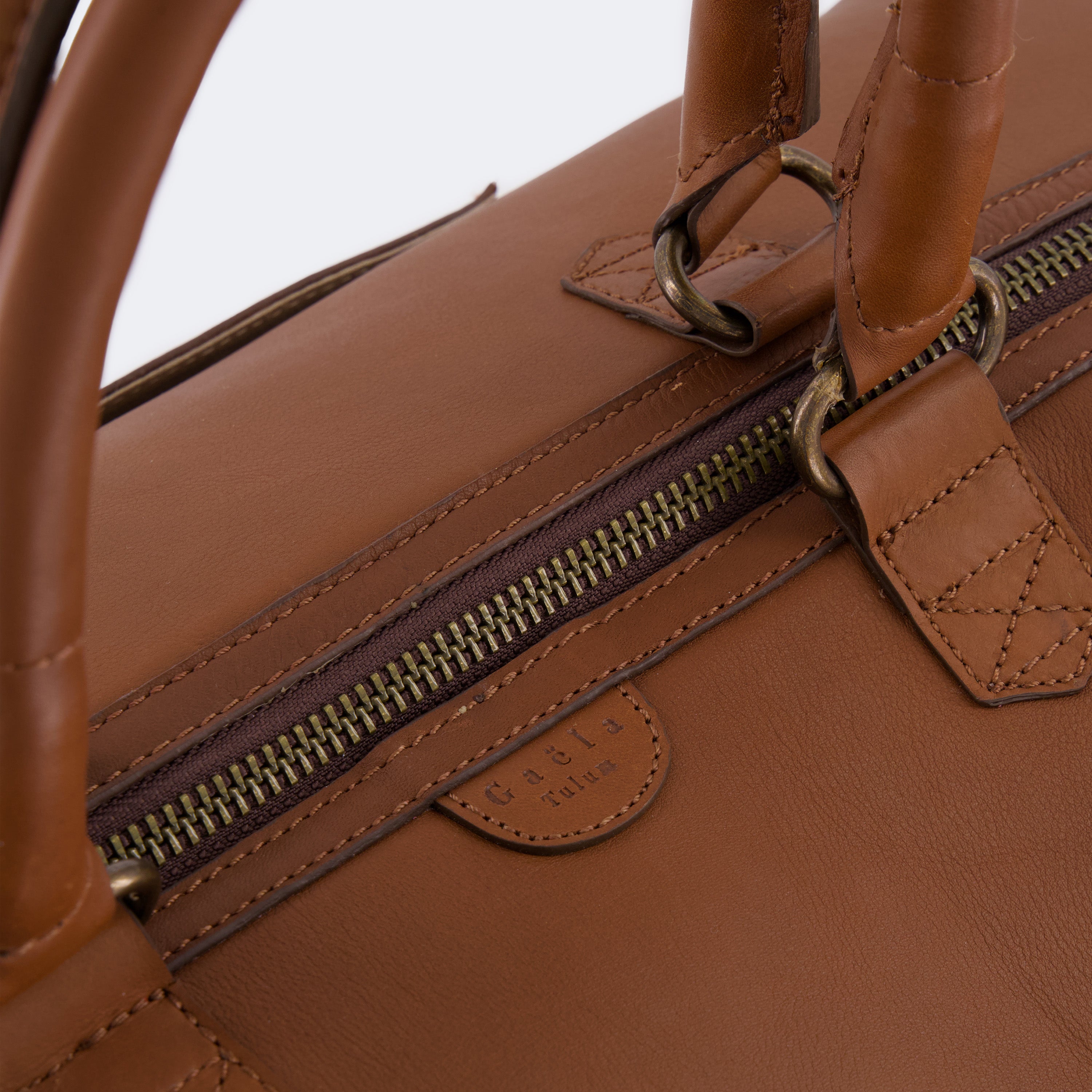 Close-up of The Valise in brown leather, showing the curved leather handles, premium hardware, and refined grain texture of the leather.
